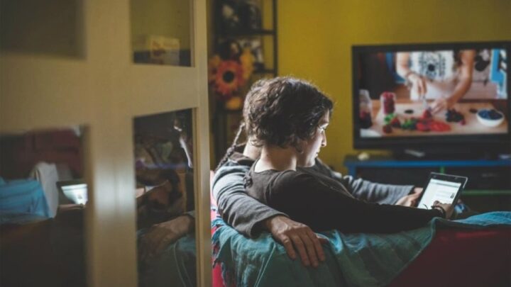 Rapaz e rapariga, no sofá, a usar um tablet, com a televisão ligada em segundo plano