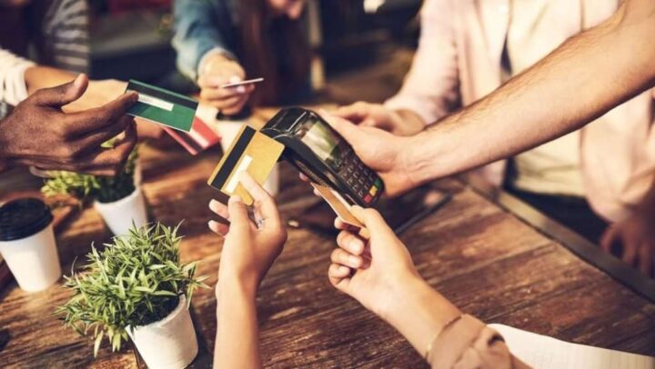 Grupo de pessoas a esticar um cartão de débito. No centro da mesa, uma máquina para pagamento com cartão