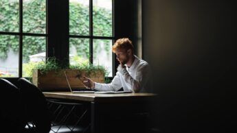 Trabalhador, no canto de uma sala, à janela, com um computador e um smartphone